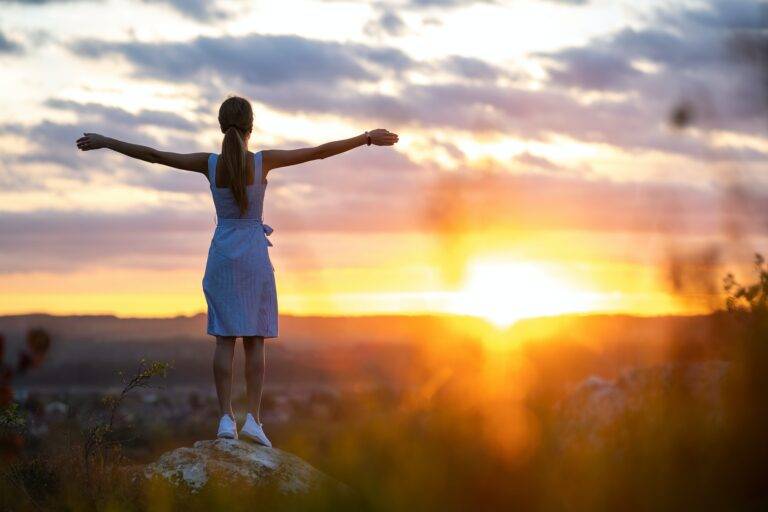 A young woman in summer dress standing outdoors with outstretched arms enjoying view of bright